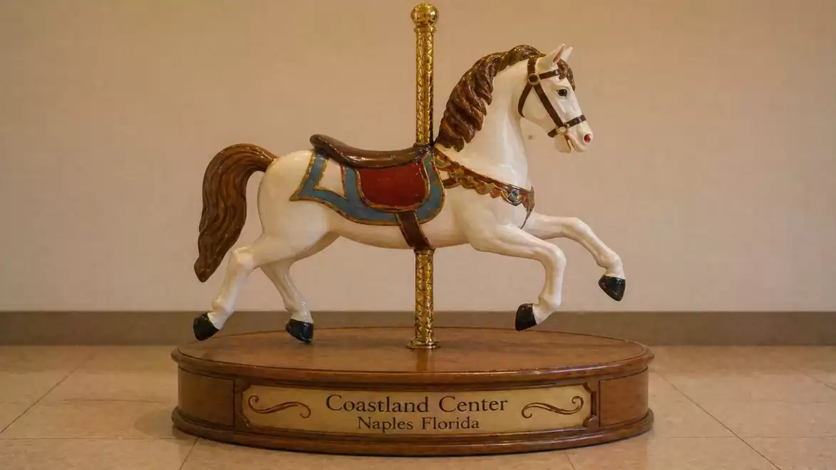 A decorative carousel horse, painted in white and brown, stands on a round wooden platform with the inscription 'Coastland Center Naples Florida' in an indoor space.