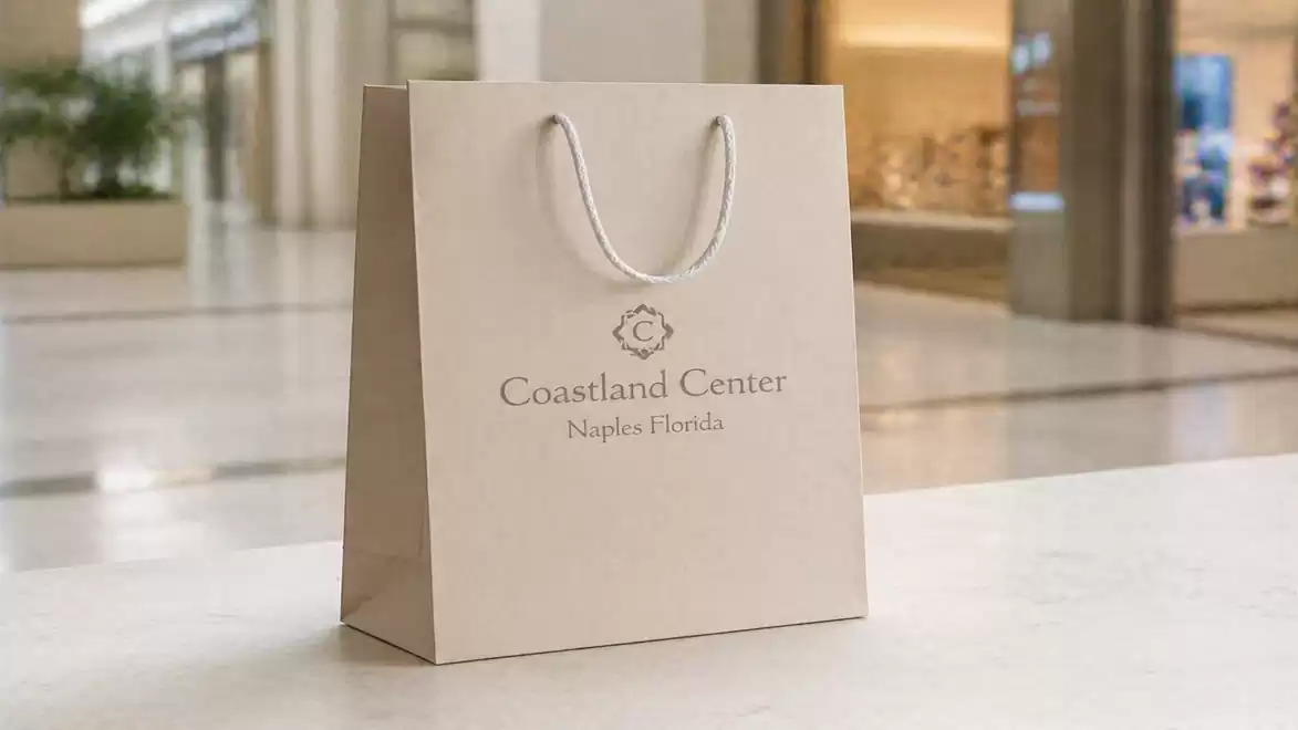 A sleek paper shopping bag featuring the words 'Coastland Center Naples Florida' stands on a light marble floor in a modern shopping mall.