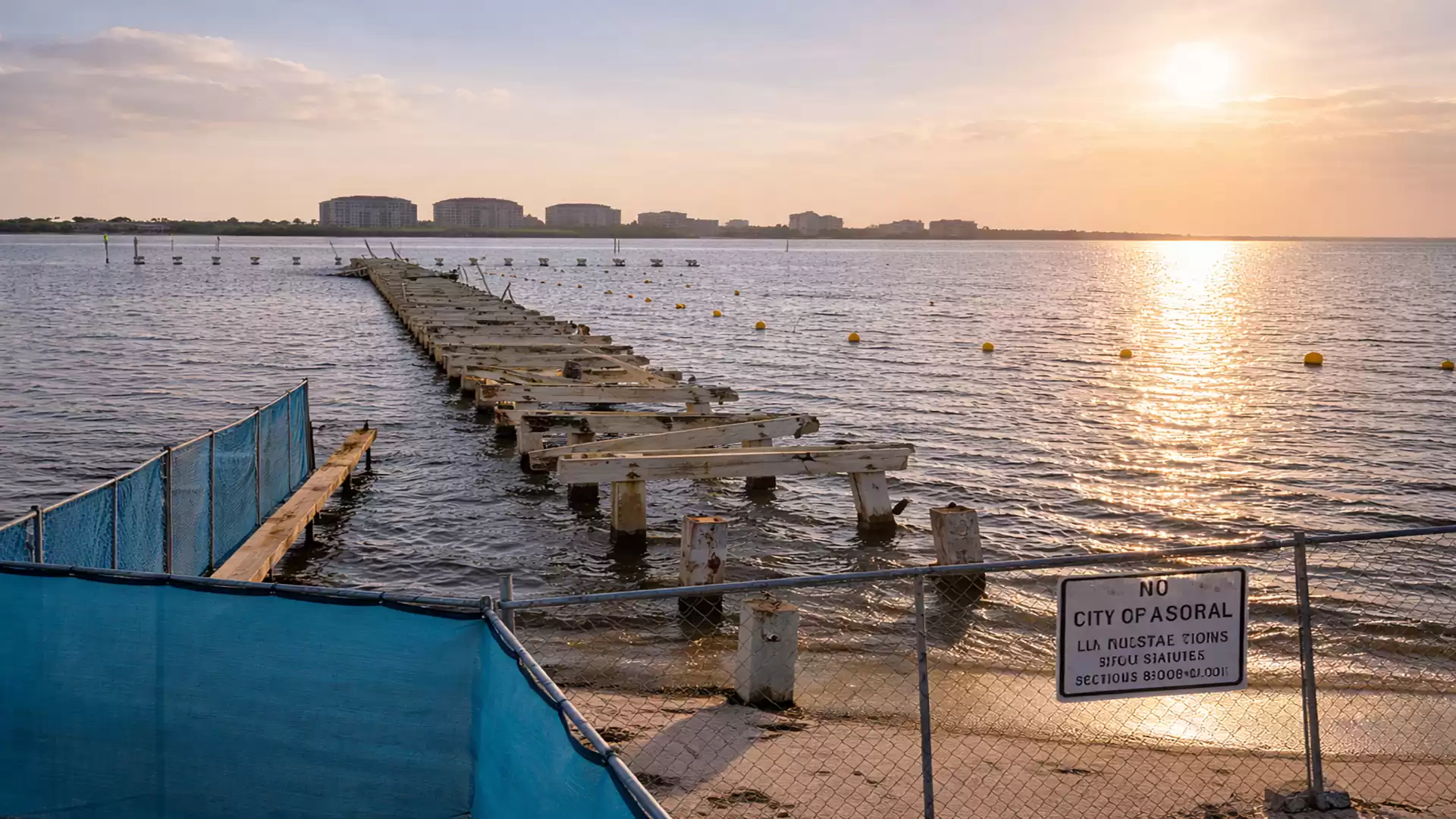An old, decaying pier leads into the water, surrounded by sunset and calm sea. A sign with text in the foreground.