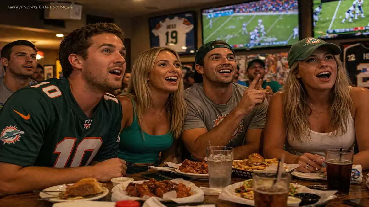 A group of young people in a sports café watching an American football game on large TV screens. They appear excited and focused as they enthusiastically watch the screens. There are plates of snacks and drinks on the table in front of them.