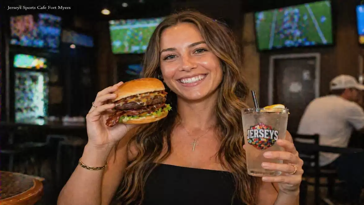 Woman holding a large burger and a drink in a sports bar, with screens showing sports broadcasts in the background.