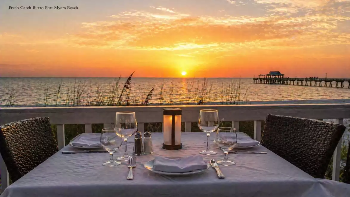An elegantly set table with white tablecloths and glasses in the foreground of a stunning ocean sunset, with a pier leading into the water.