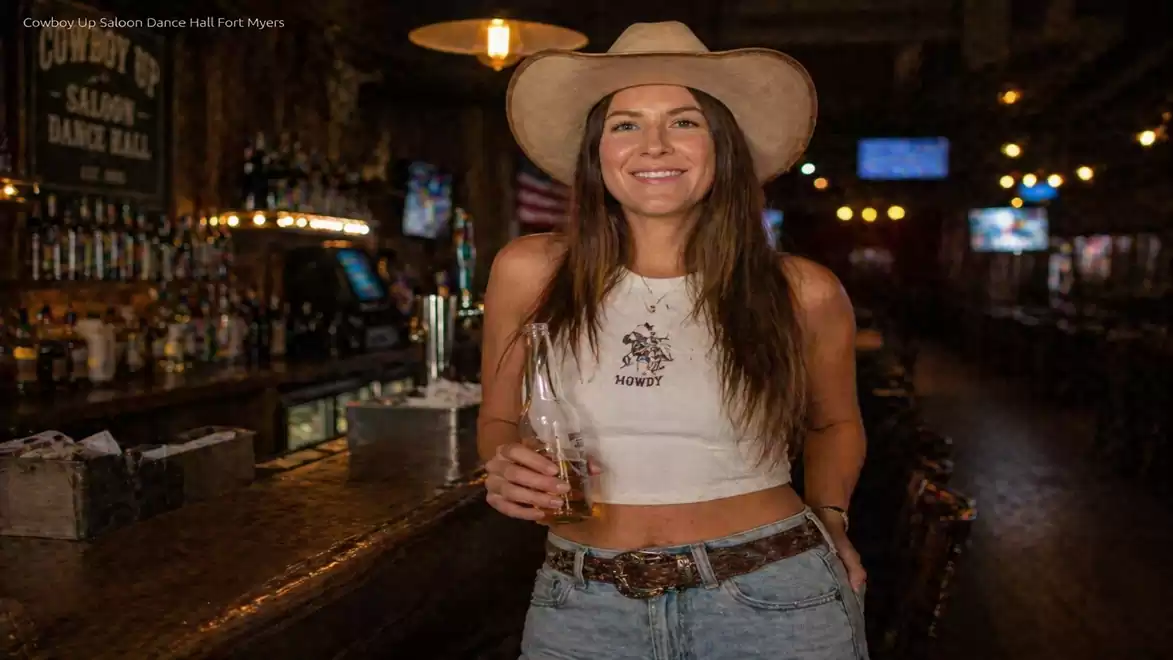 Woman wearing a cowboy hat and crop top stands in a dimly lit bar holding a drink.