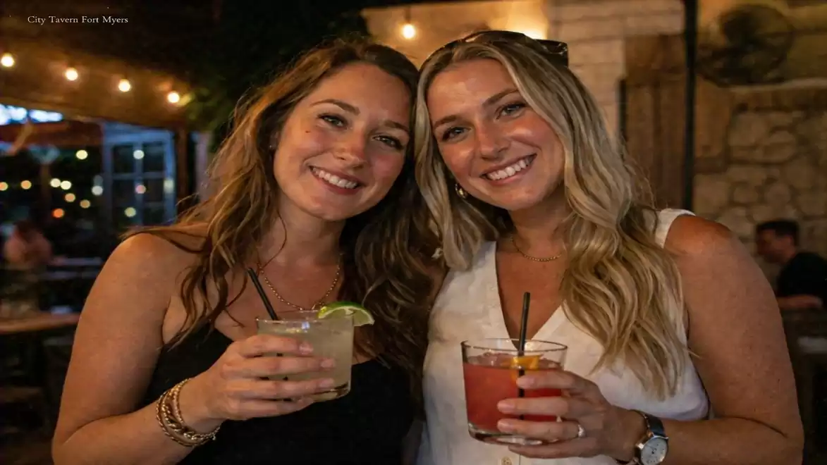 Two women smiling with cocktails in their hands in a dimly lit bar.