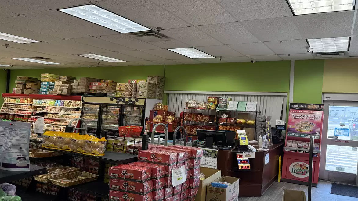 A small supermarket with shelves full of various food products and a checkout area. Beverage coolers are visible in the background.