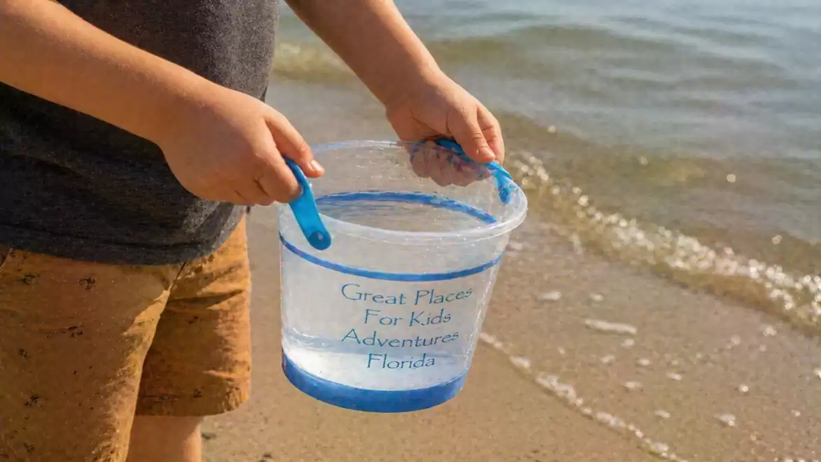 A child holds a water-filled bucket at the beach as gentle waves roll onto the sand.