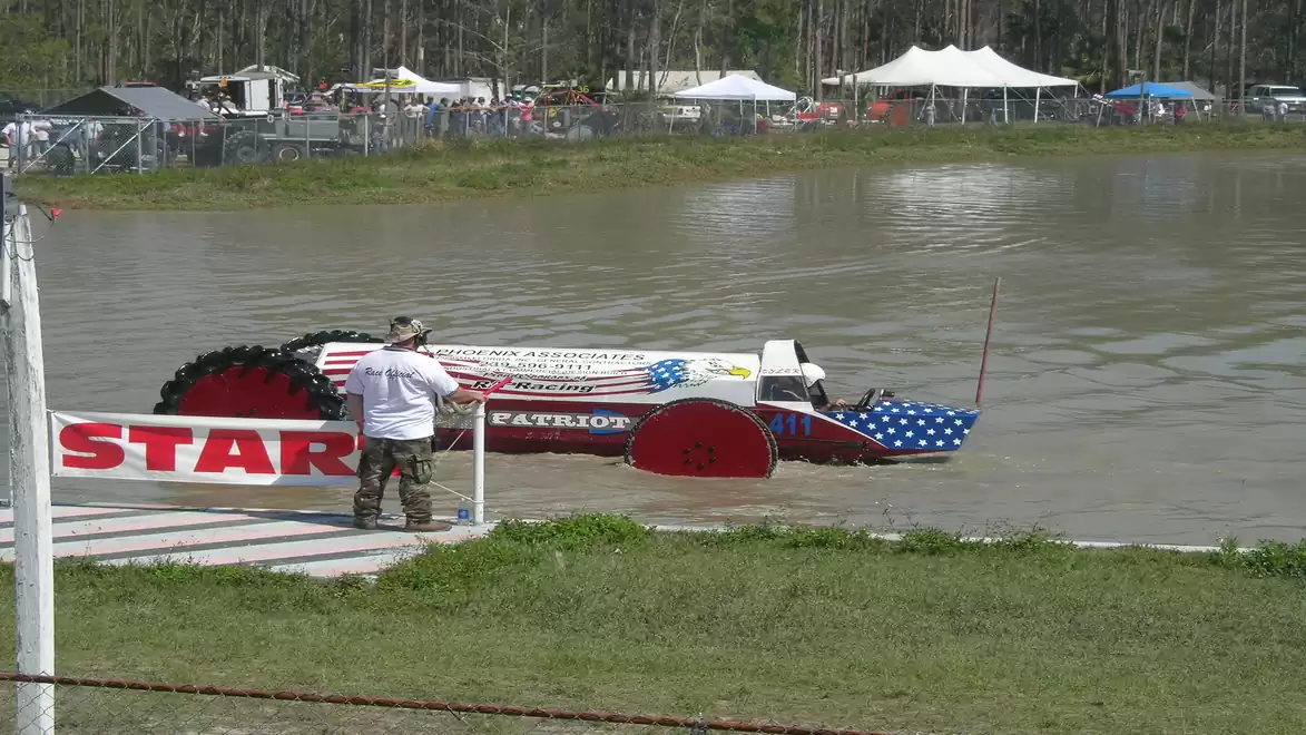 An amphibious vehicle with an American flag design readies for the water start of a race, surrounded by a park with tents in the background.