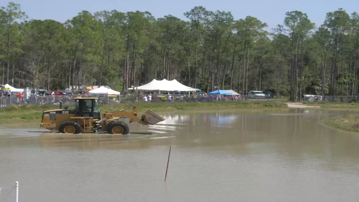 A yellow excavator is in a large, muddy water pool, surrounded by trees and white tents in the background.
