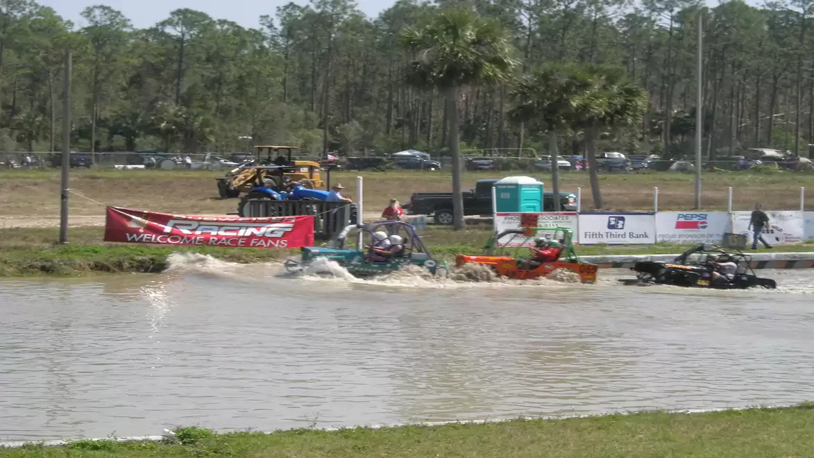 Men drive modified lawn mowers through a water trench during an outdoor race, with spectators watching in the background.