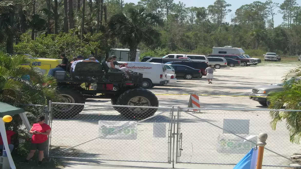 A large monster truck is parked in a parking lot, surrounded by several cars and a person walking by. Trees and a sunny sky are visible in the background.