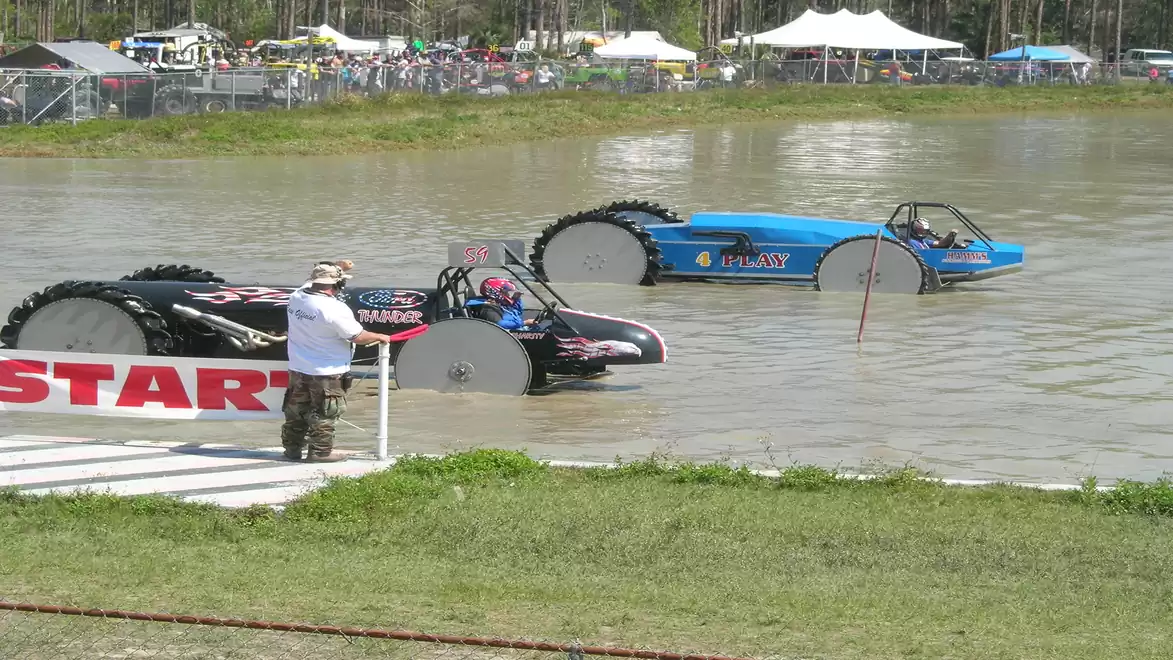 Two vehicles with large wheels prepare for a mud buggy race in a water pit, while a man in a hat stands at the start.