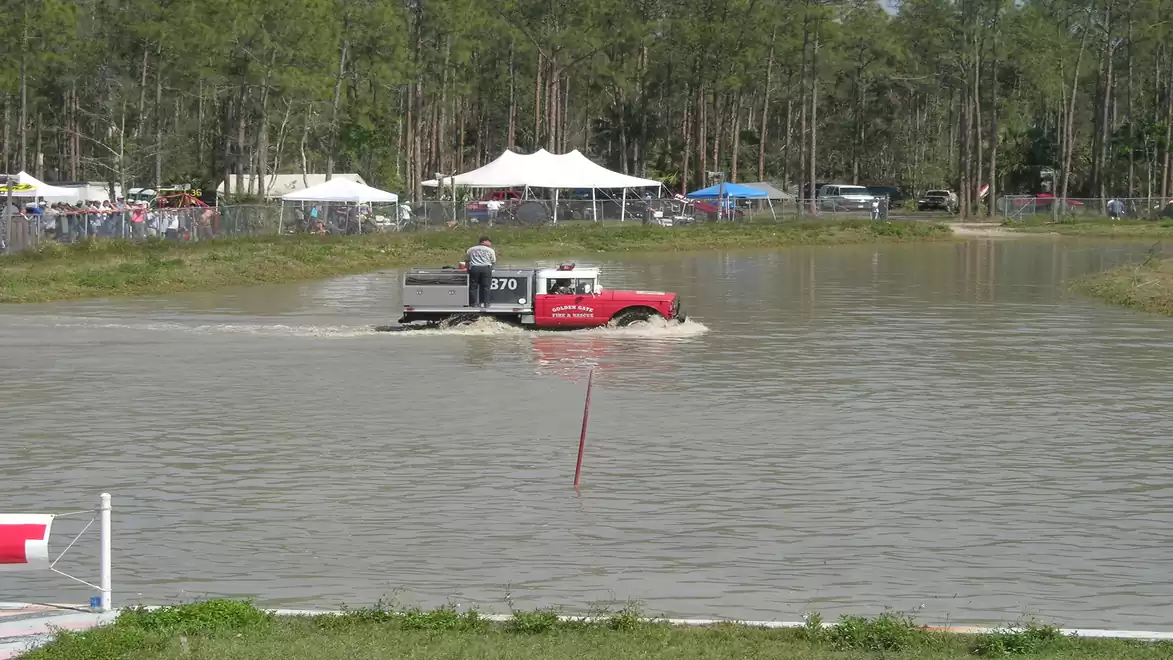 A red off-road vehicle drives through a large muddy water ditch in a rural setting with trees and tents in the background.