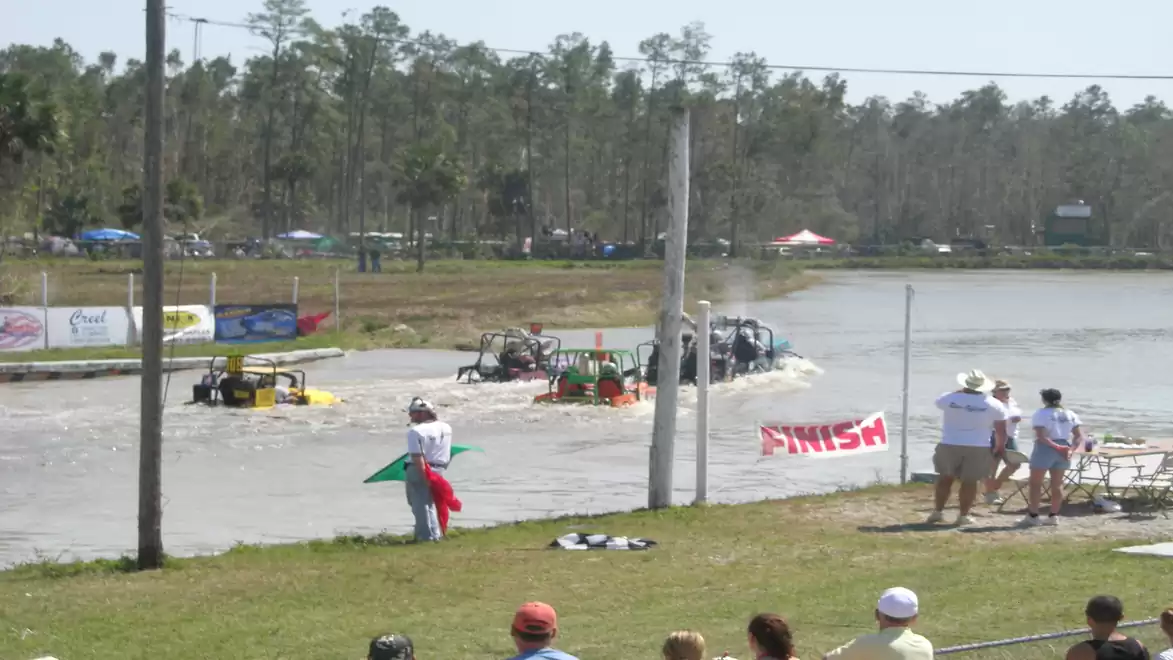Off-road vehicle race in water, where several 4x4s navigate a muddy course with spectators watching and a flagger standing nearby.