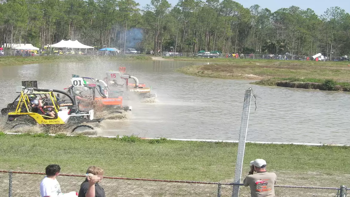 Various off-road vehicles drive through a muddy water hole at an outdoor event, with spectators watching.