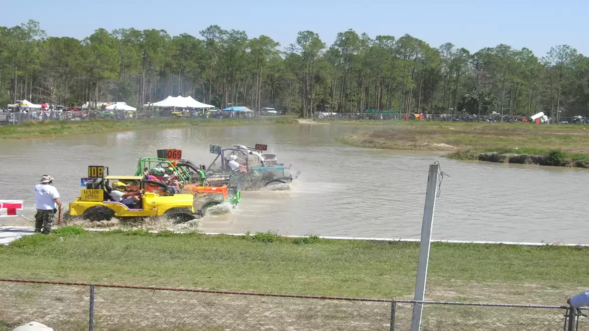 Off-road vehicles race through a muddy water course during an off-road event, surrounded by a green forest backdrop.
