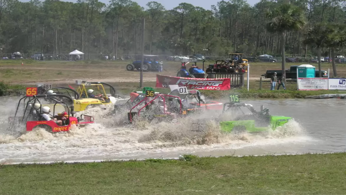 Several off-road vehicles cross a body of water during a race, surrounded by a dusty landscape with trees in the background.