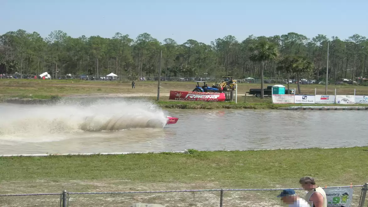 A red vehicle drives through a water ditch at an offroad event, with a spectator standing in the foreground.