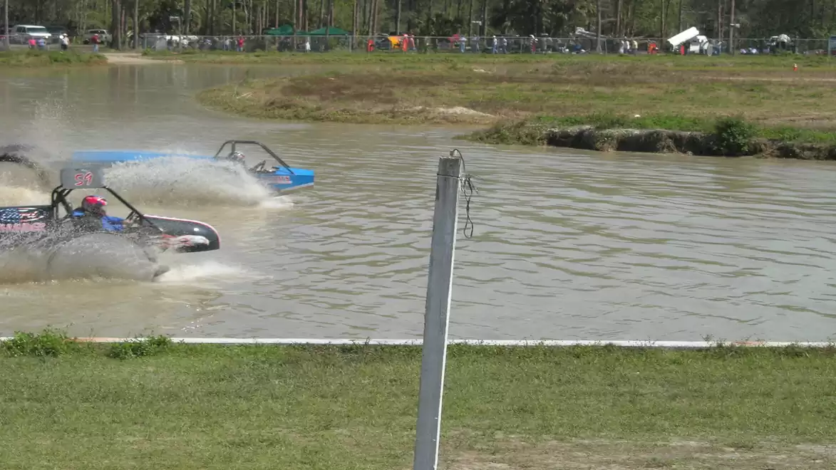 Two off-road vehicles drive through a water hole at an off-road event. Crowds in the background watch the action.