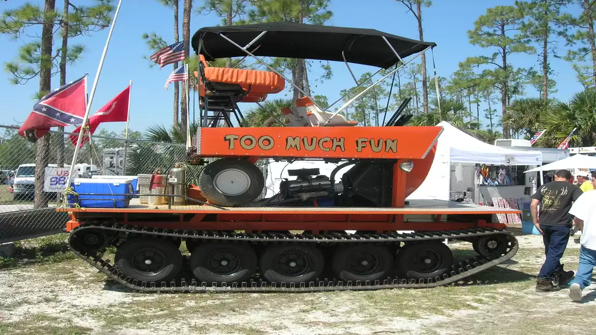 An orange tank vehicle with the inscription 'Too Much Fun' is parked on grassy ground under a blue sky at an event. Tents, people, and trees are visible in the background.