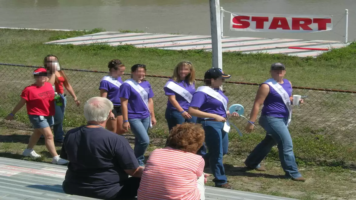 Group of people at an outdoor event, some wearing sashes near a starting line.