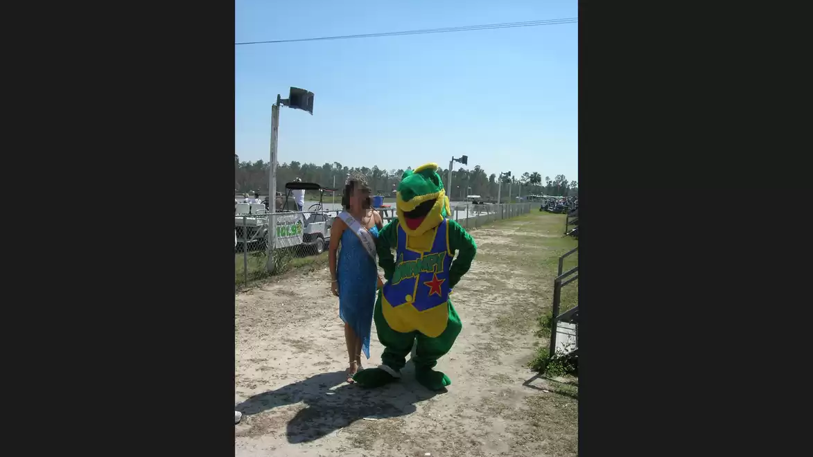 A person in a blue dress poses next to a large green mascot in a colorful costume on a dusty path in sunny weather.