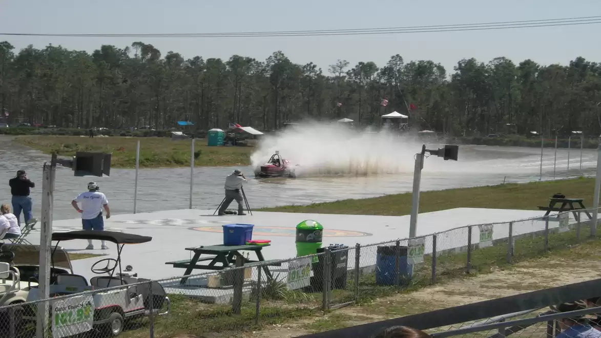 A racing boat speeds across a water track as spectators watch from behind a fence.