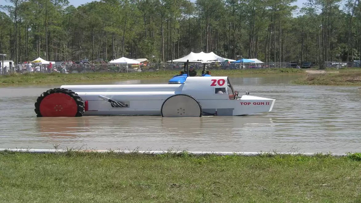 A tractor drives through a muddy, watered area on a field. Trees and tents are visible in the background.