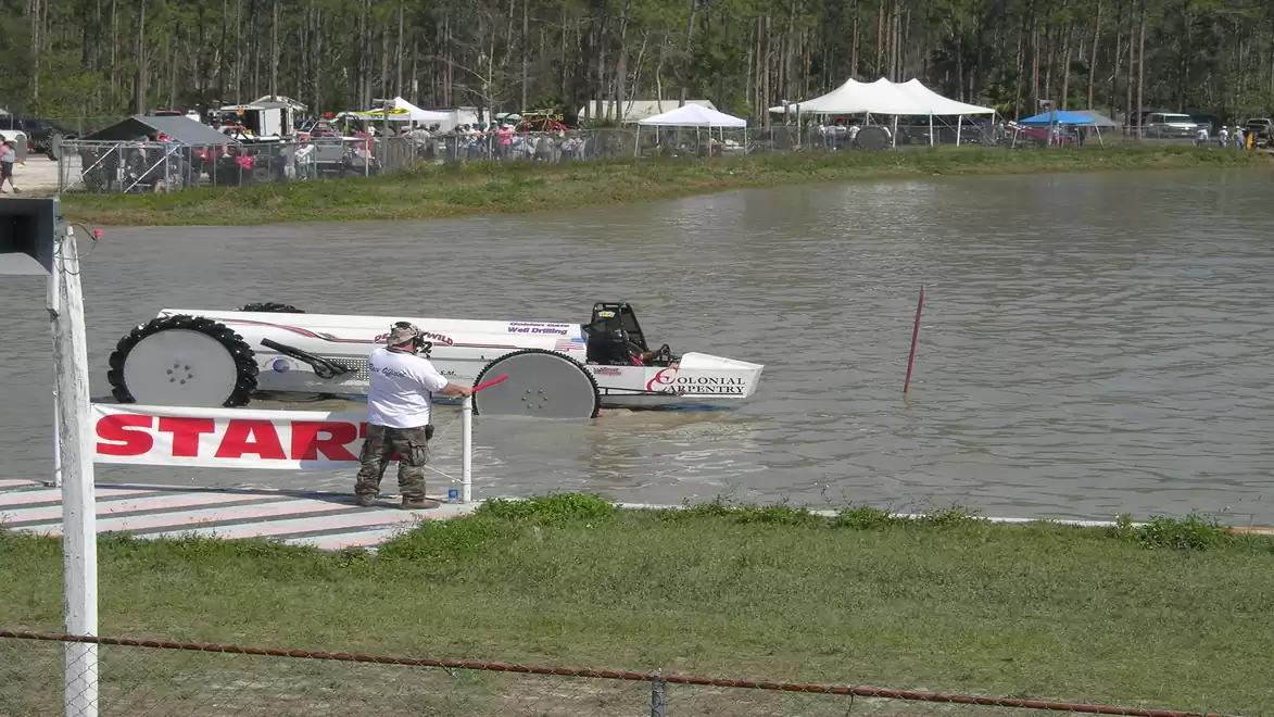 A modified vehicle enters the water at a tractor racing event with a mud track. Spectators and tents in the background.