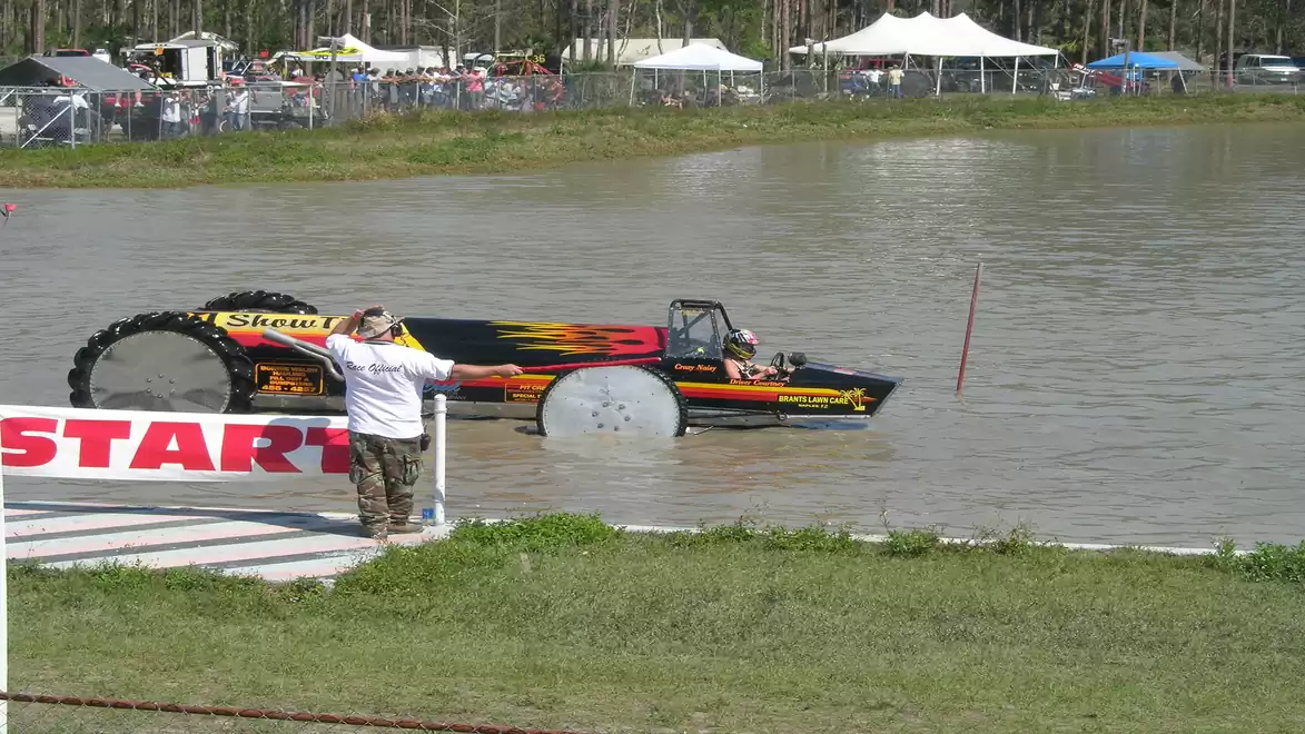 Tractor with large wheels driving across a lake at an outdoor event with spectators.