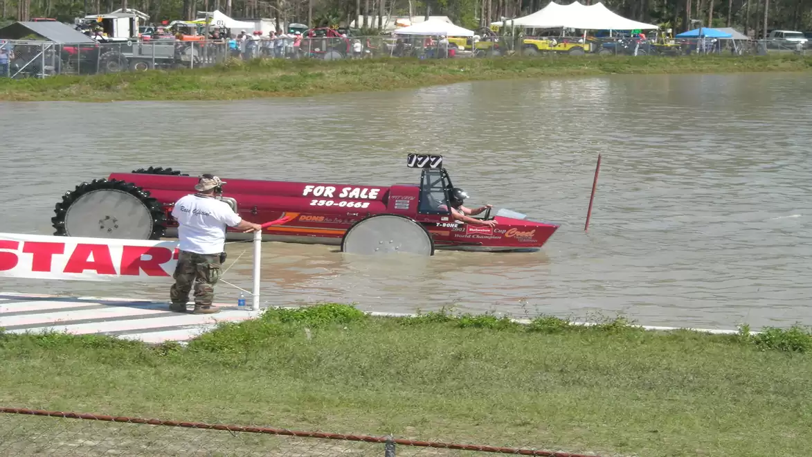A red vehicle partially submerged in water at an event with several people in the background.