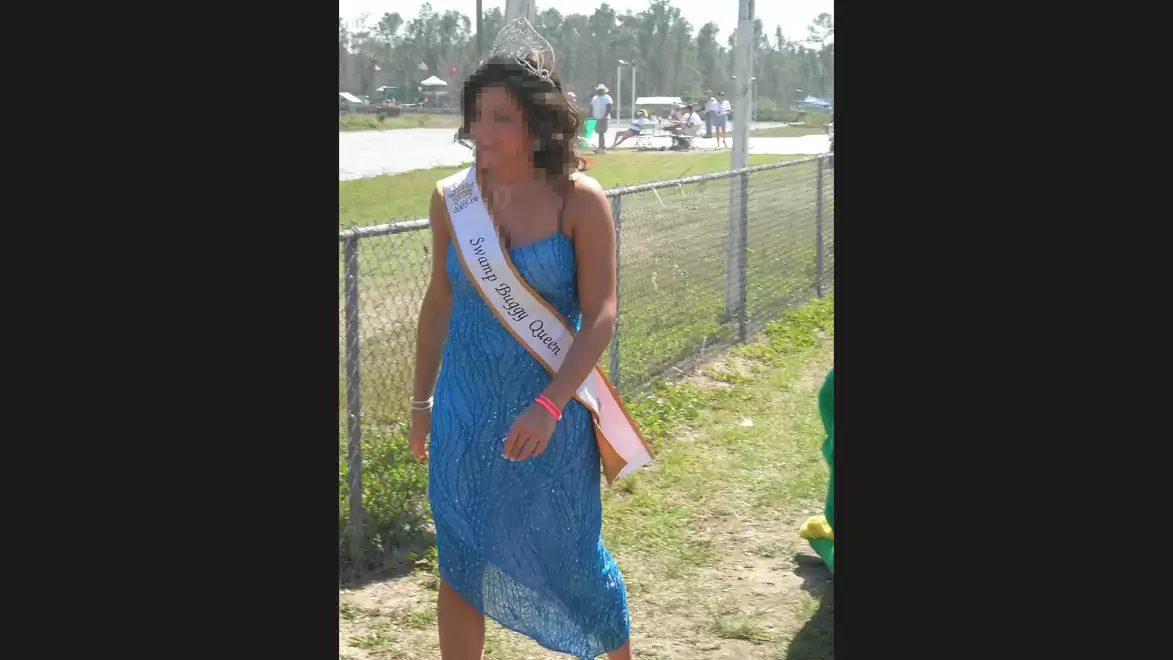 Woman in a blue dress with a sash stands next to a fence in sunny weather.