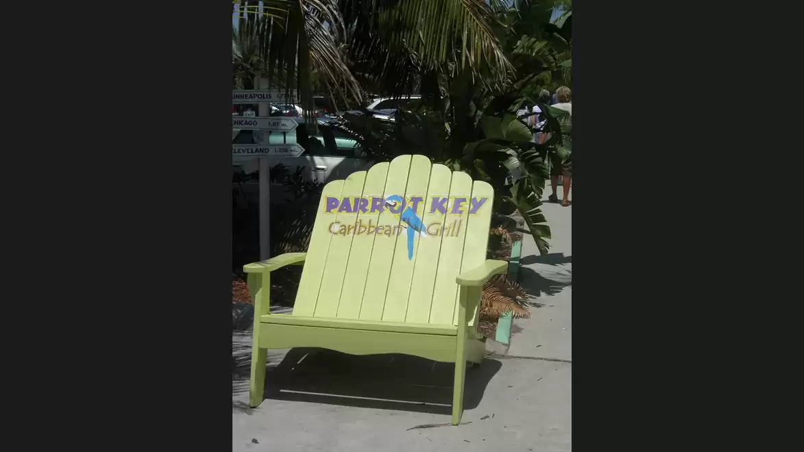 A large yellow wooden chair is situated outside on a sunny patio next to green palm trees. The backrest of the chair features the logo of 'Parrot Key Caribbean Grill'.