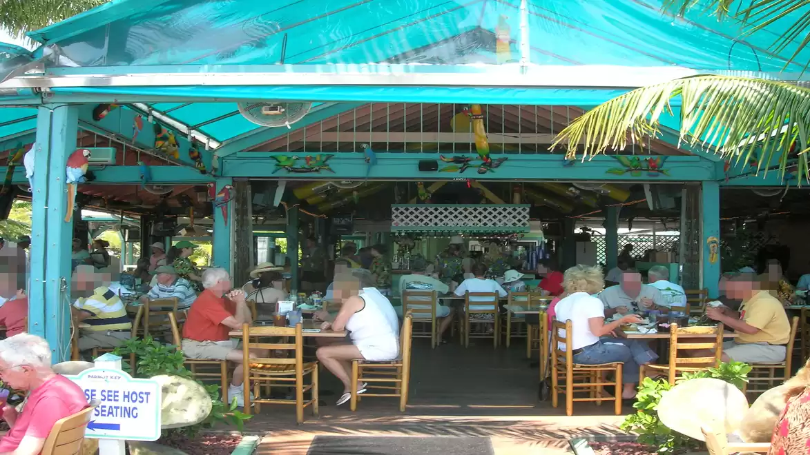 A bustling outdoor restaurant with several guests dining under a turquoise roof. The setting feels tropical with palm trees and colorful decorations.