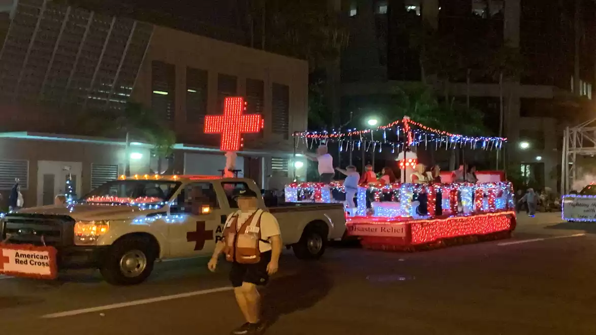 An illuminated American Red Cross parade float adorned with lights and a large red cross moves through the streets in a nighttime parade.