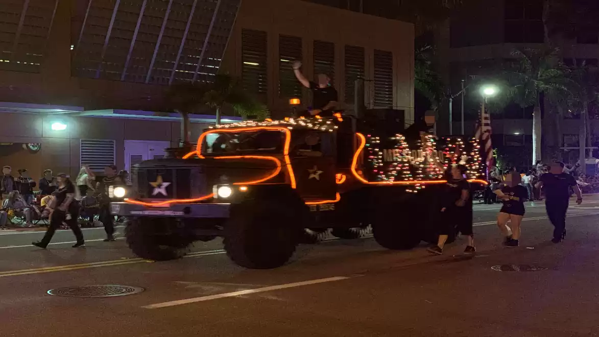A festively decorated truck with string lights participates in a nighttime street parade, with people lining the street.