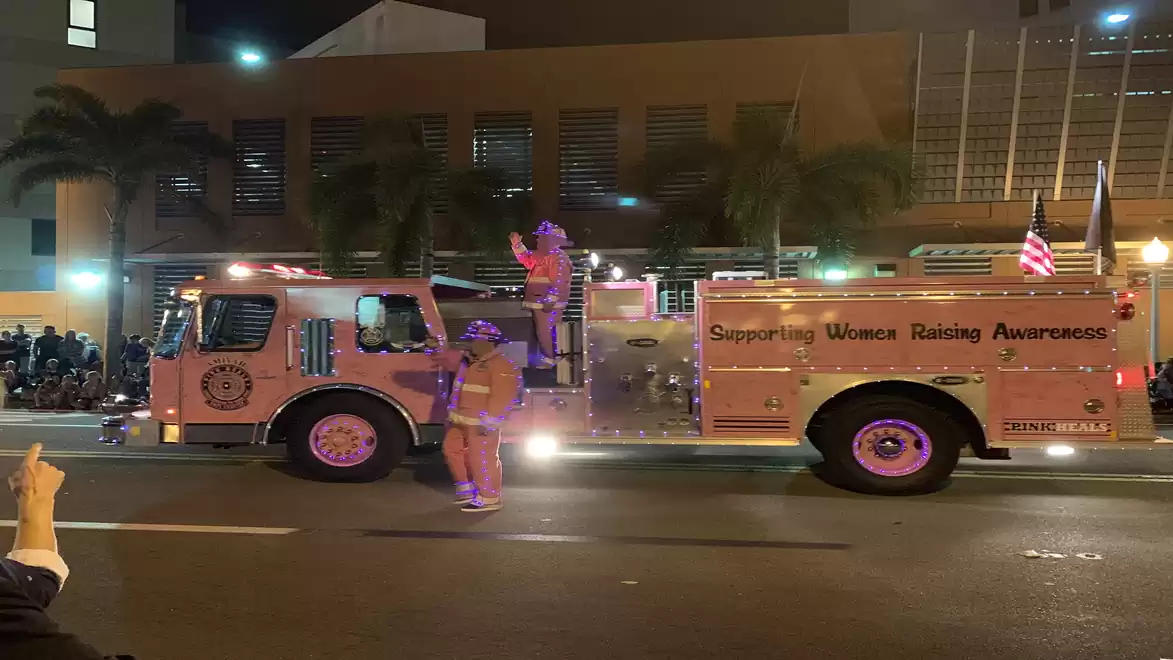 A fire truck with the text 'Supporting Women Racing Awareness' drives through a city at night with illuminated buildings in the background. People are wearing lights and costumes.