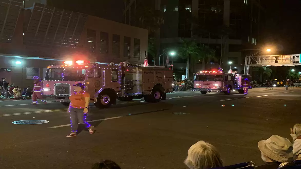 Fire trucks at night parade on illuminated street, people watching.