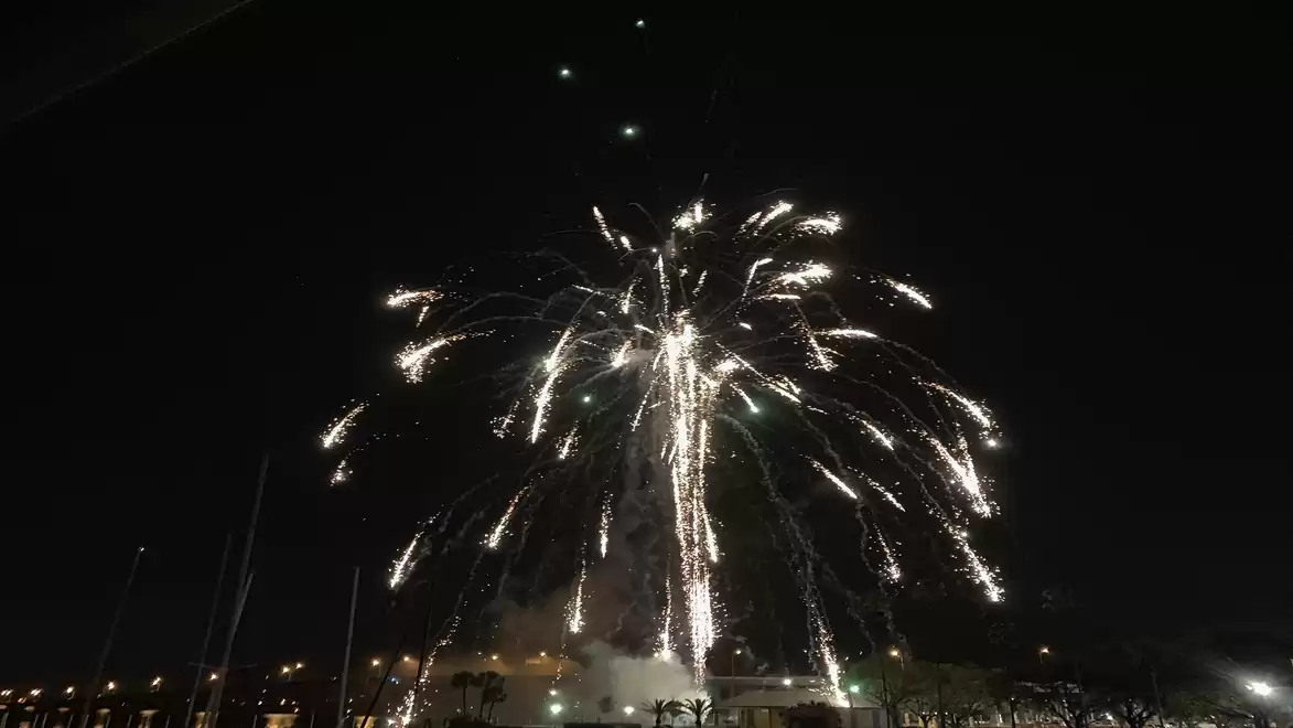 A large fireworks display lights up the night sky over a city, with clear white and golden trails impressively appearing above the buildings.