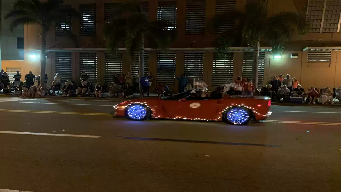 A lit-up red car with blue wheel lights drives past a crowd at night.