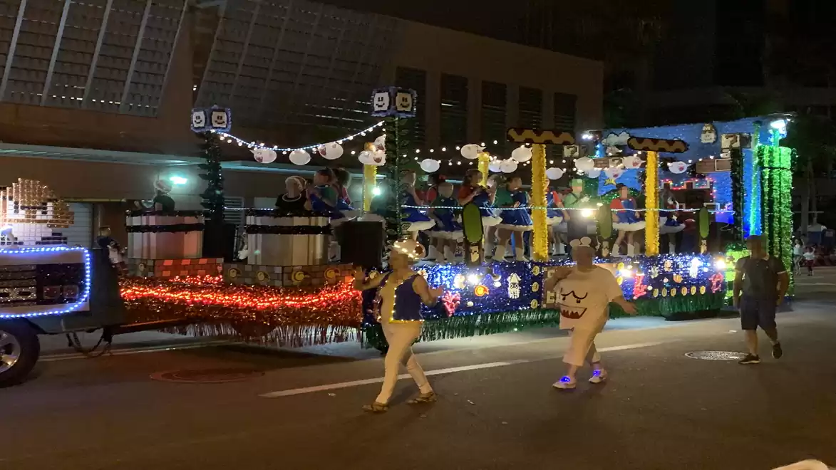 Festively lit carnival float driving down a street at night, decorated with colorful lights and paper lanterns, accompanied by costumed individuals.