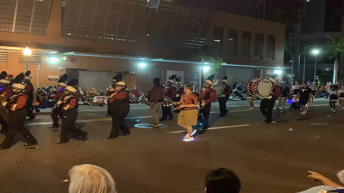Street scene at night with a marching band in uniforms playing drums and other instruments.