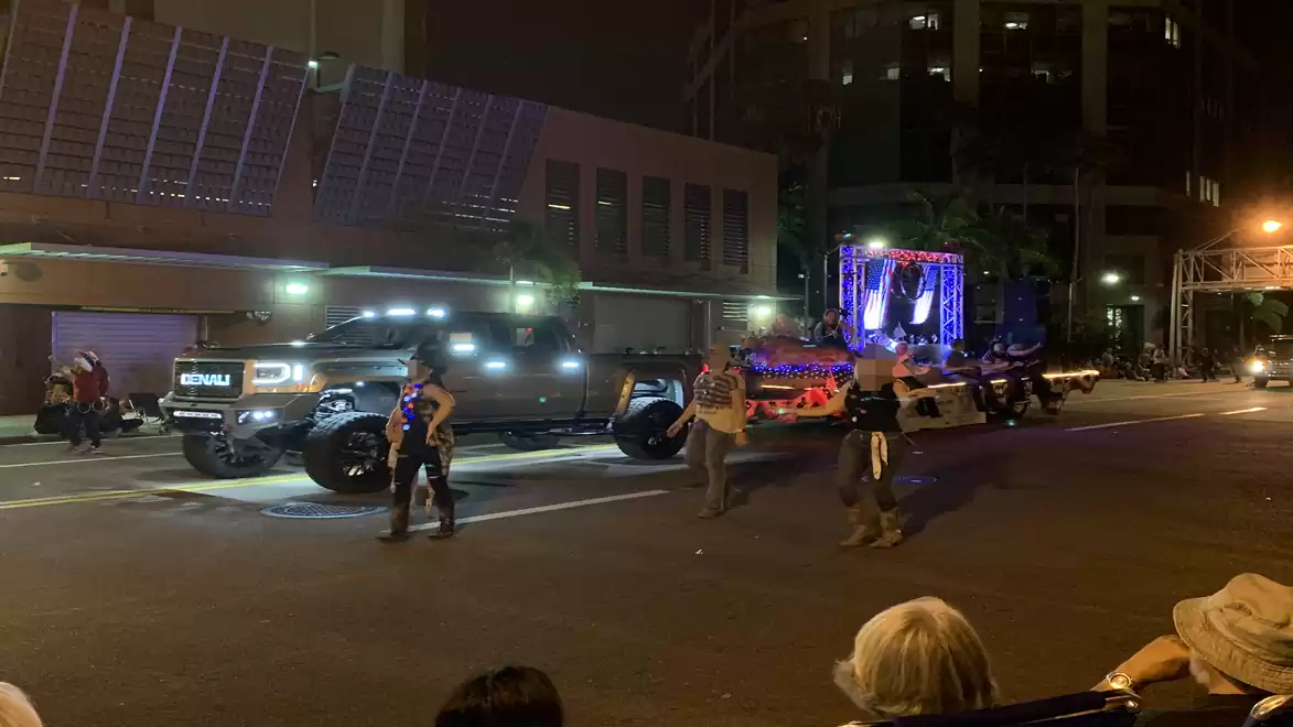 A nighttime parade with a large illuminated truck, followed by a festively decorated trailer, moving through a city street. People are walking alongside the truck.