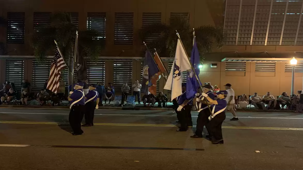 A group of people in uniform carry various flags during a parade on a nighttime street.