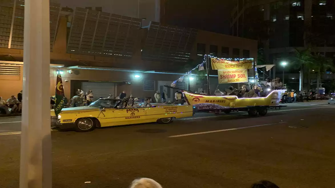 Night parade on a city street with a long yellow car towing a brightly lit platform with people and decorations.