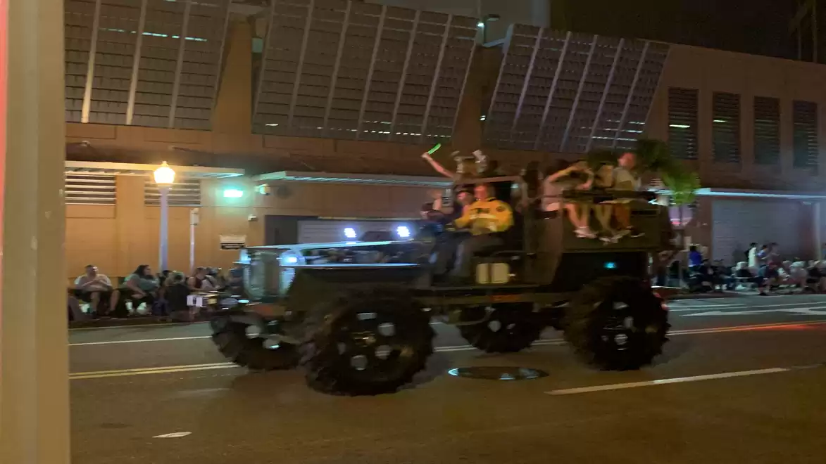Night scene of a parade with a large, illuminated vehicle carrying people driving on the street, while spectators sit at the roadside.