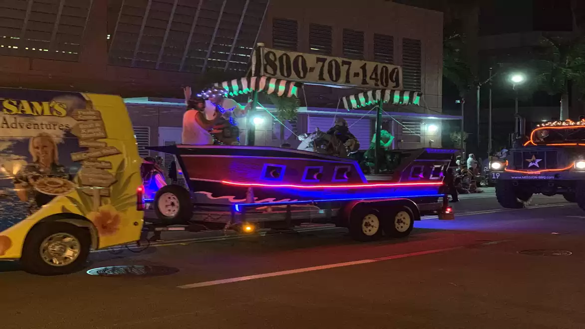 Nighttime photo of a festive parade with brightly lit vehicles and trailers in vibrant colors.