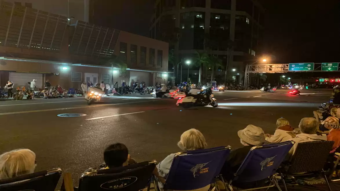 Nighttime image of a motorcycle parade on a city street, with spectators in folding chairs and illuminated buildings in the background.