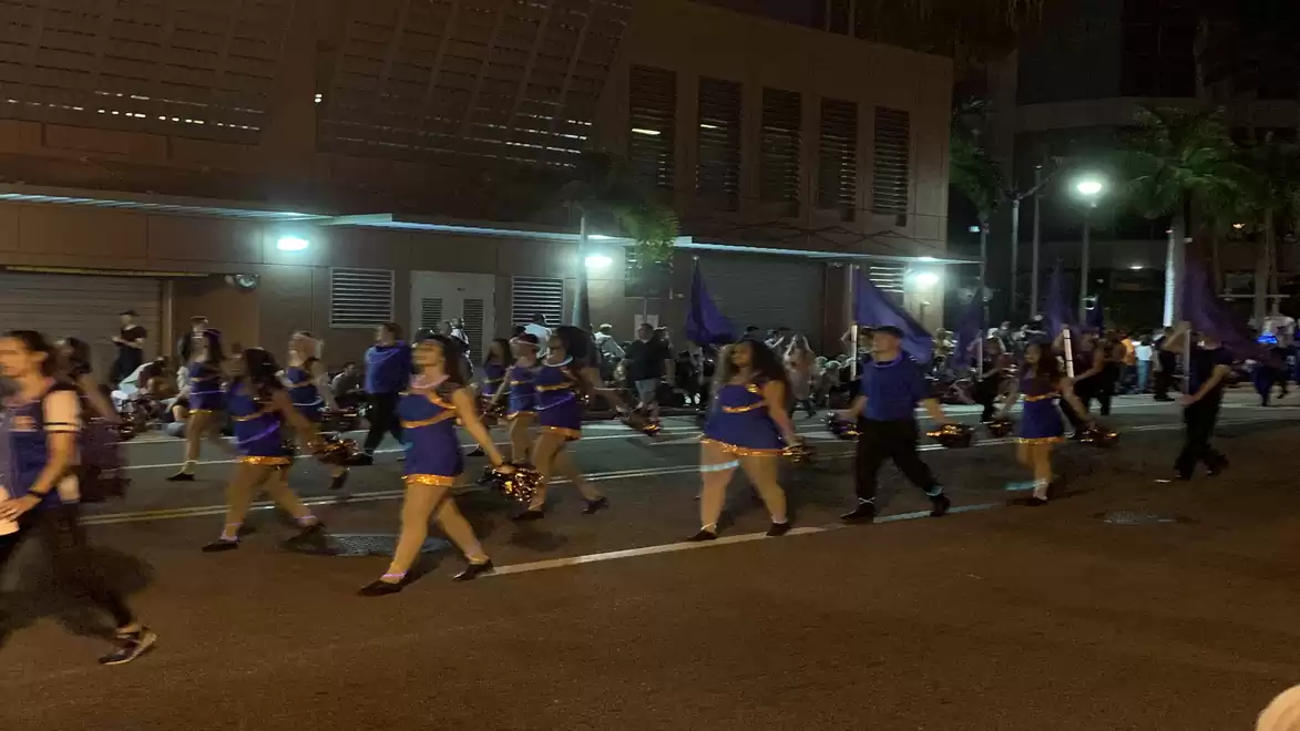 A nighttime parade with people in blue outfits marching on a lit sidewalk.