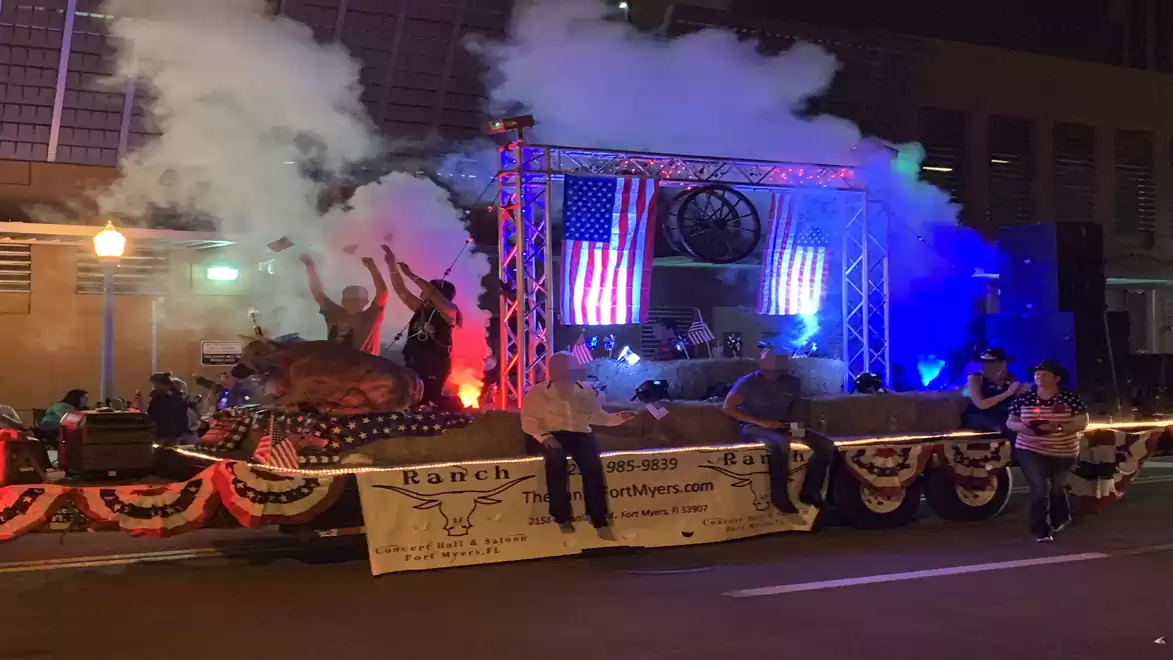 An illuminated parade float at night featuring a stage where a band is performing. American flags and colorful smoke clouds enhance the scene.