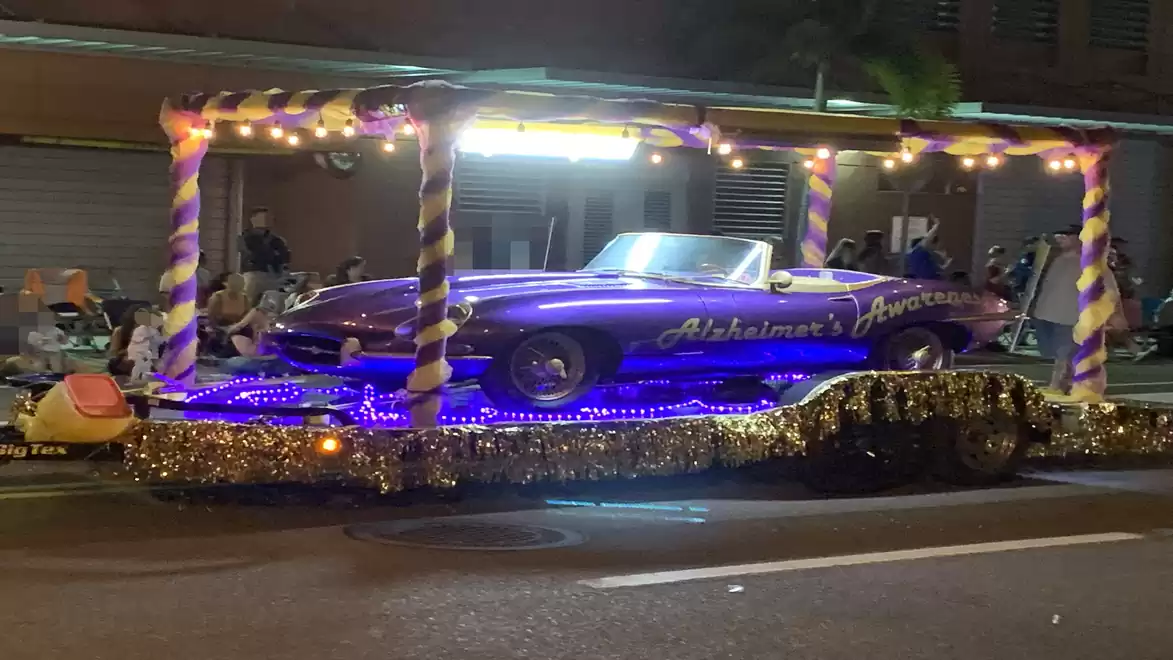 A purple convertible is displayed on a festively decorated float at a parade. The float is adorned with lights and glitter, while spectators sit by the roadside.
